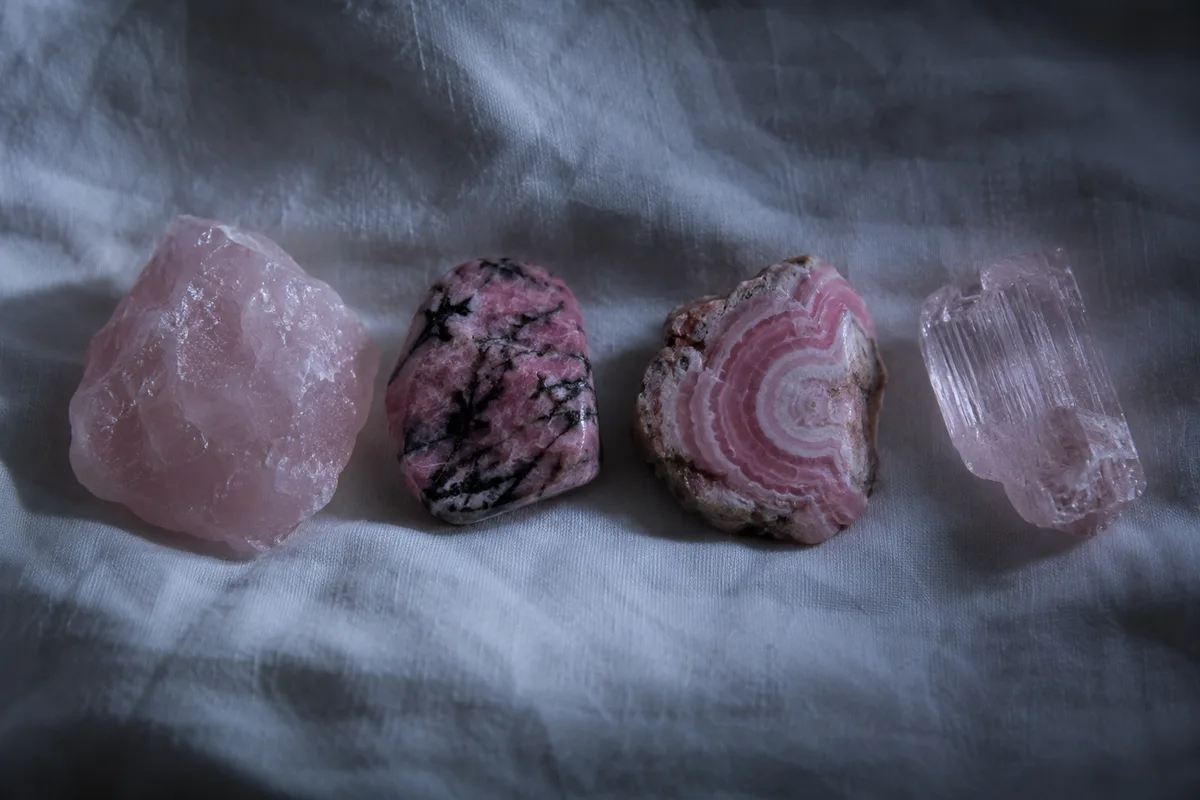Quatre pierres roses alignées sur un linge de coton blanc, quartz rose, rhodonite veinée de noir, rhodochrosite rubanée et kunzite claire